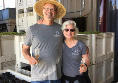 Bruce Lundquist and Penny Gadd-Coster, executive director of winemaking, standing in front of stacked harvest grape bins with glasses of sparkling wine glasses in their hands.