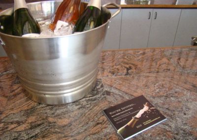 Large wine chiller bucket with 3 bottles of Rack & Riddle sparkling wine on ice sitting on a counter next to “you’re invited” postcards.