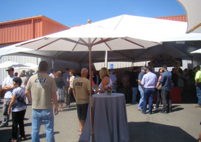 Guests at the grand opening event talking to each other under market umbrellas and a tent.