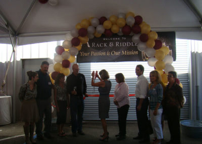 Ribbon cutting ceremony at our 2014 Grand Opening celebration at our Healdsburg, California facility. Rebecca Faust using giant scissors to cut the red ribbon on our new Healdsburg Facility in 2014. Bruce Lundquist is standing next to her along with members of the Healdsburg Chamber of Commerce board. There is a balloon arch in the background.